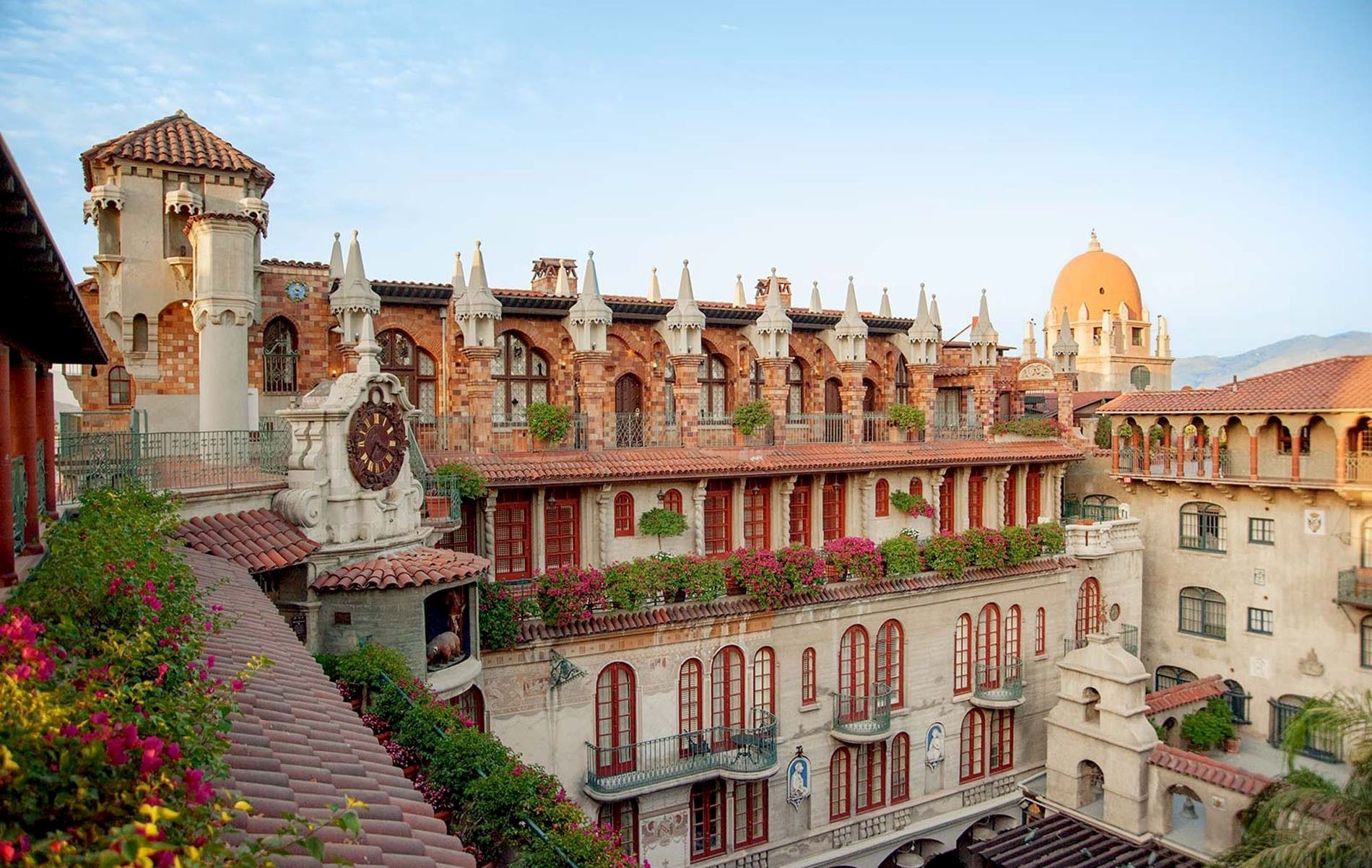 Spanish-style resort courtyard with red-tiled roofs, arched windows, a domed tower, and colorful flowers under a blue sky.
